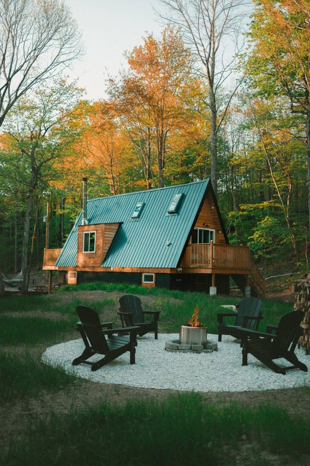 A large A-frame house with a teal metal roof and wooden siding sits in a golden autumn forest, featuring a foreground fire pit area with black Adirondack chairs on white gravel.
