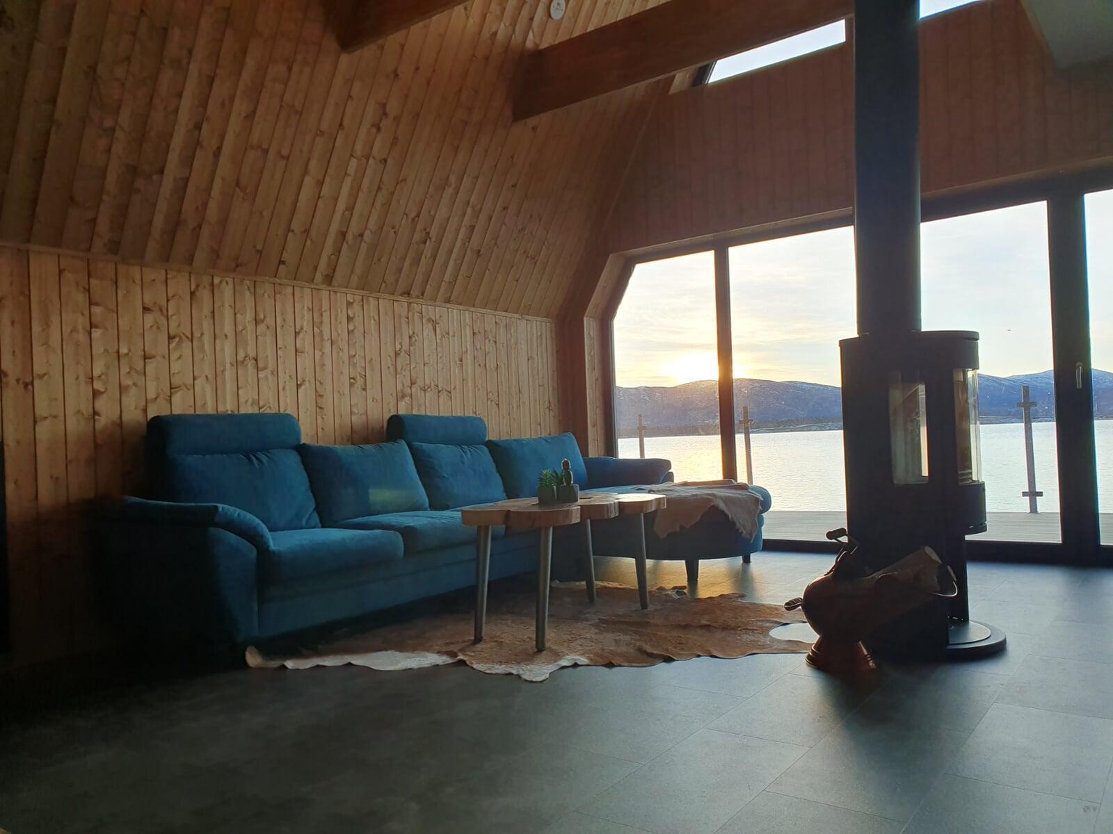 A cozy living room inside an A-frame cabin featuring a blue sectional sofa, a wood-burning stove, and a large window overlooking a scenic lake at sunset.