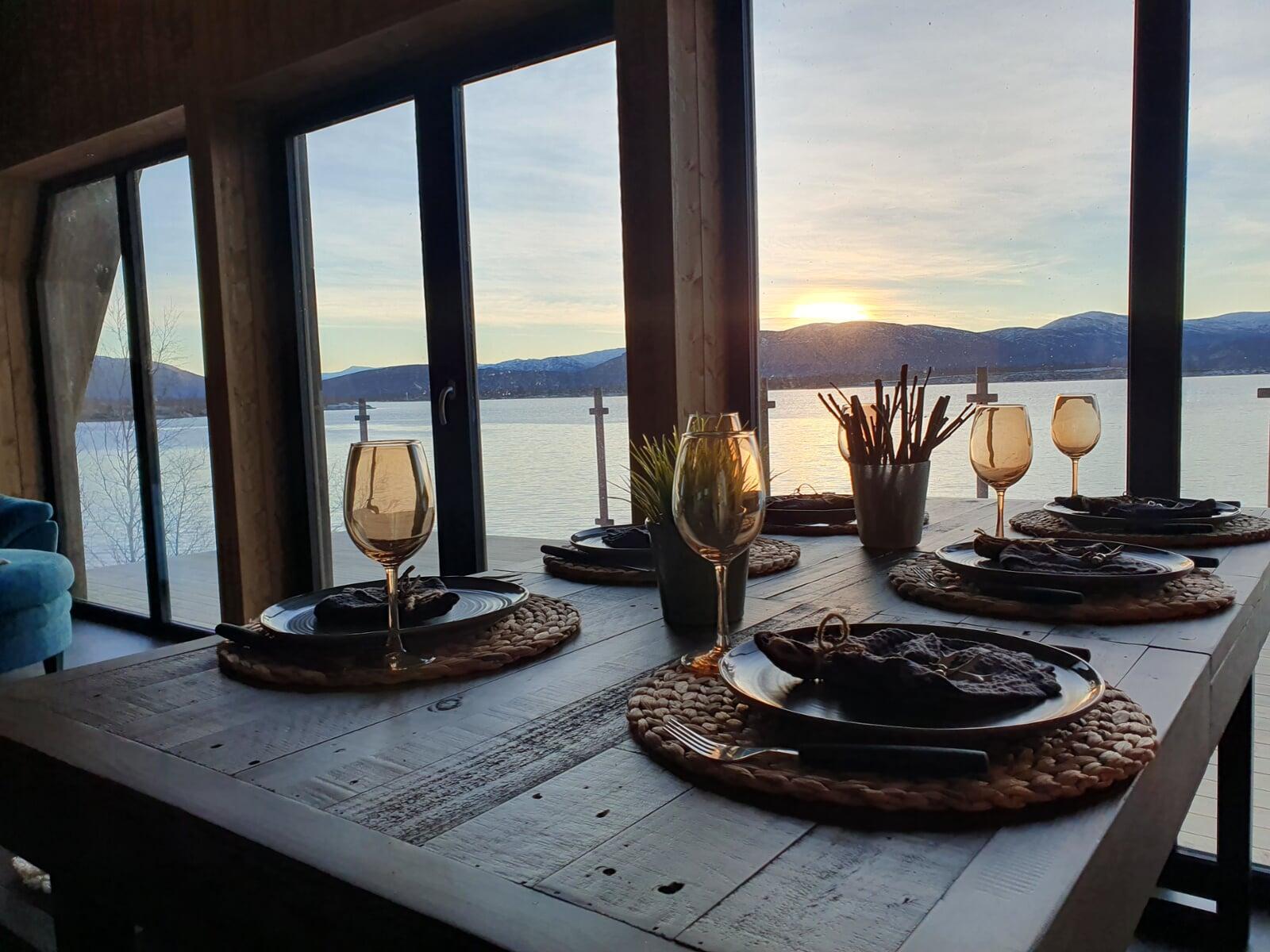 An elegantly set wooden dining table with woven placemats and wine glasses faces a large window, offering a stunning view of a sunset over a calm lake and distant mountains.