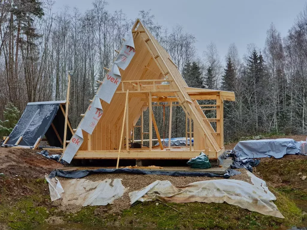 The wooden frame of an A-frame cabin is under construction on a snowy lot, with partially applied protective building wrap visible on the angled roof structure.