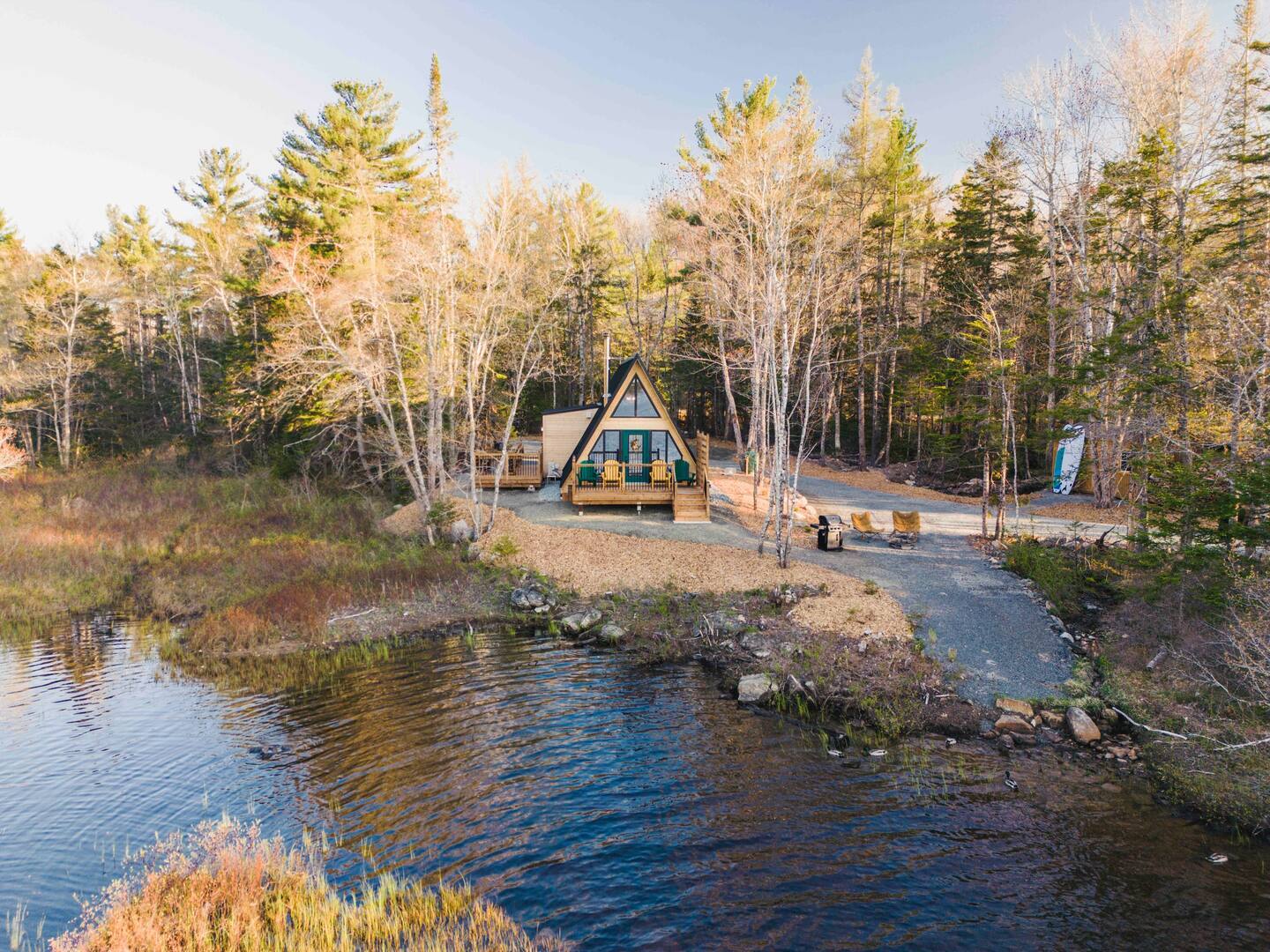 A charming A-frame cabin with a wooden front deck and yellow chairs sits at the edge of a calm lake, surrounded by a dense forest of evergreen and birch trees under a clear sky.