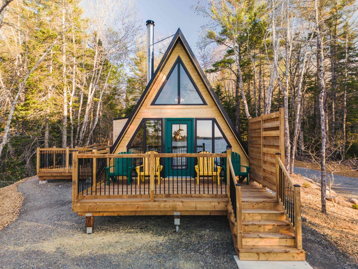 A front-facing view of a light-wood A-frame cabin with a teal front door, featuring a raised wooden deck with yellow and green chairs nestled in a serene forest setting.