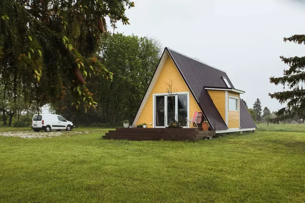 A bright yellow A-frame house with a dark roof and a small wooden deck sits on a green lawn, with a white van parked nearby and a line of lush trees in the background.