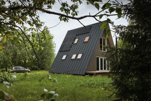 A modern A-frame cabin featuring a dark metal roof equipped with solar panels and skylights sits in a lush green meadow, framed by the branches of nearby trees.