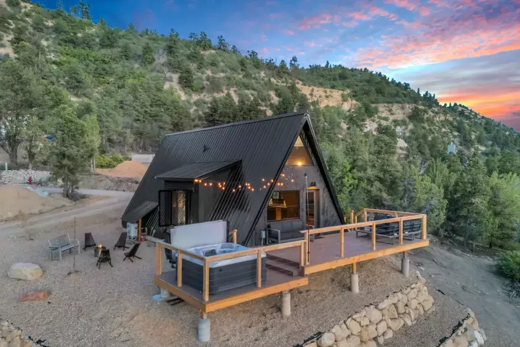 A modern black A-frame house sits on a rocky hillside at sunset, featuring an expansive wooden deck with a hot tub and string lights overlooking a forested mountain valley.