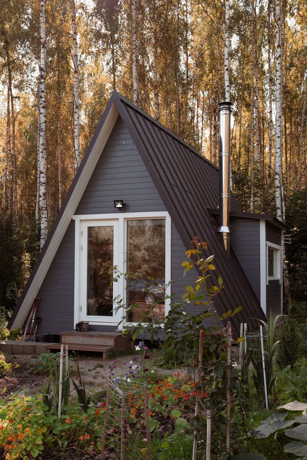 A modern grey A-frame cabin with white-trimmed doors and a tall silver chimney sits nestled in a sunlit birch forest, fronted by a lush garden with colorful flowers.
