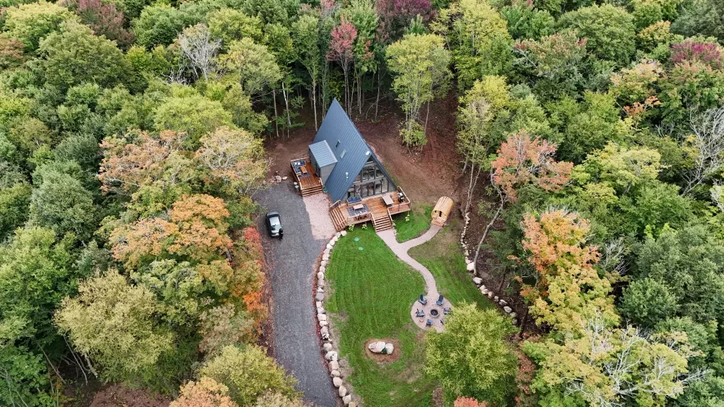 An aerial view captures a modern A-frame cabin nestled within a lush, colorful autumn forest, complete with a gravel driveway, spacious wooden deck, and a circular fire pit area on the lawn.