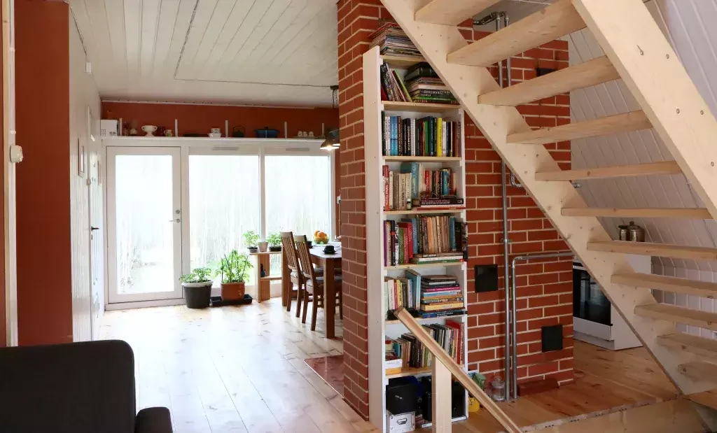 A bright, modern interior of an A-frame house features light wood floors, an open wooden staircase, and a central brick pillar with a built-in bookshelf.