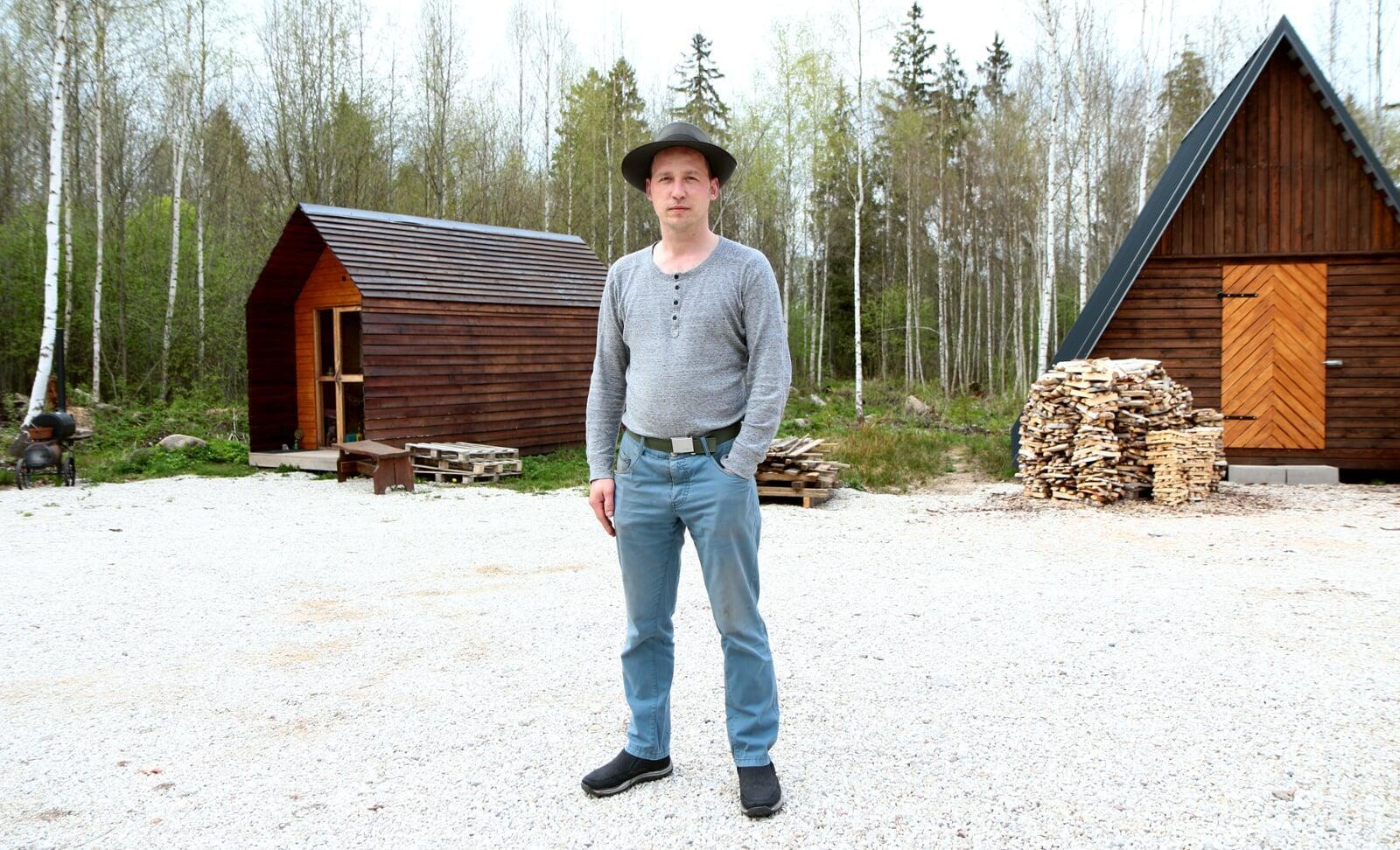 A man wearing a hat stands in a gravel clearing between a small wooden cabin and an A-frame structure with a stack of firewood nearby.