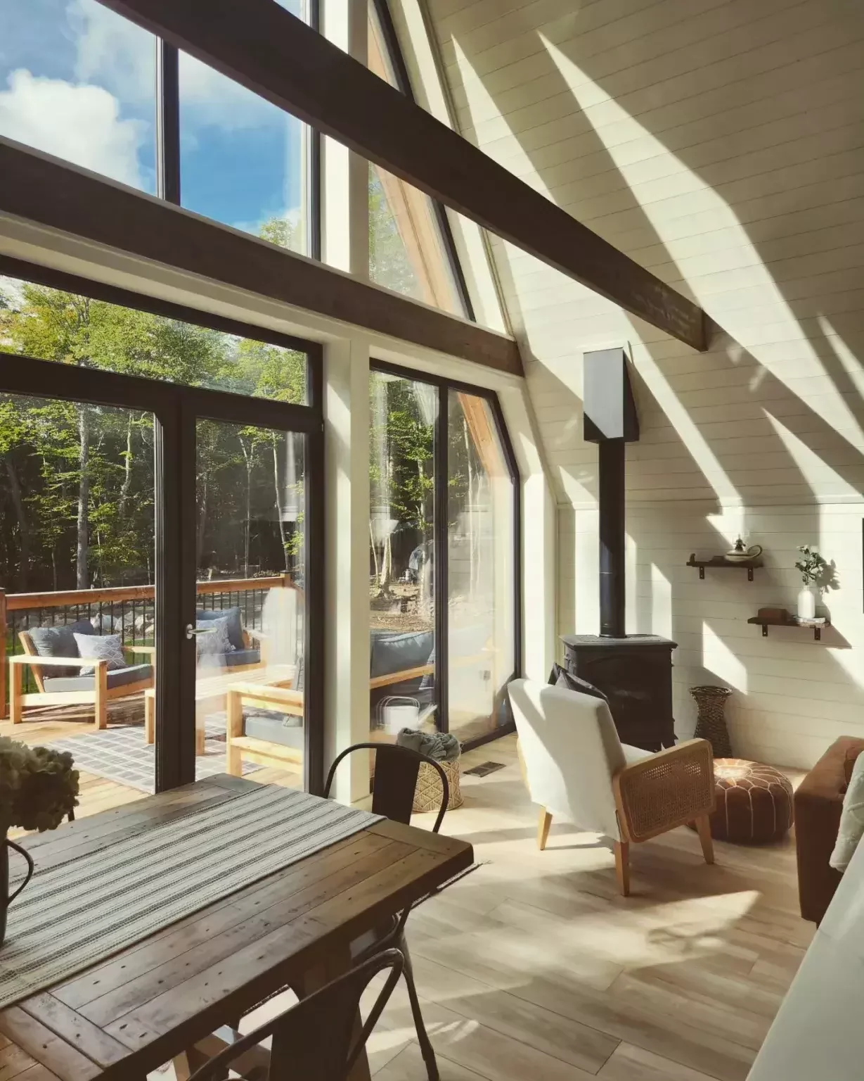 A sun-drenched A-frame cabin interior features white paneled walls, floor-to-ceiling windows, a wood-burning stove, and a dining table, creating an airy and inviting living space.