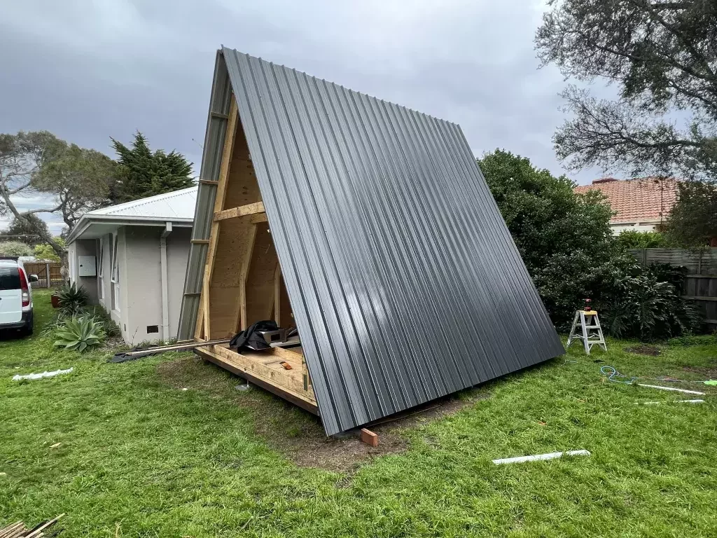 A partially completed A-frame structure with a grey metal roof stands in a backyard next to a residential house.