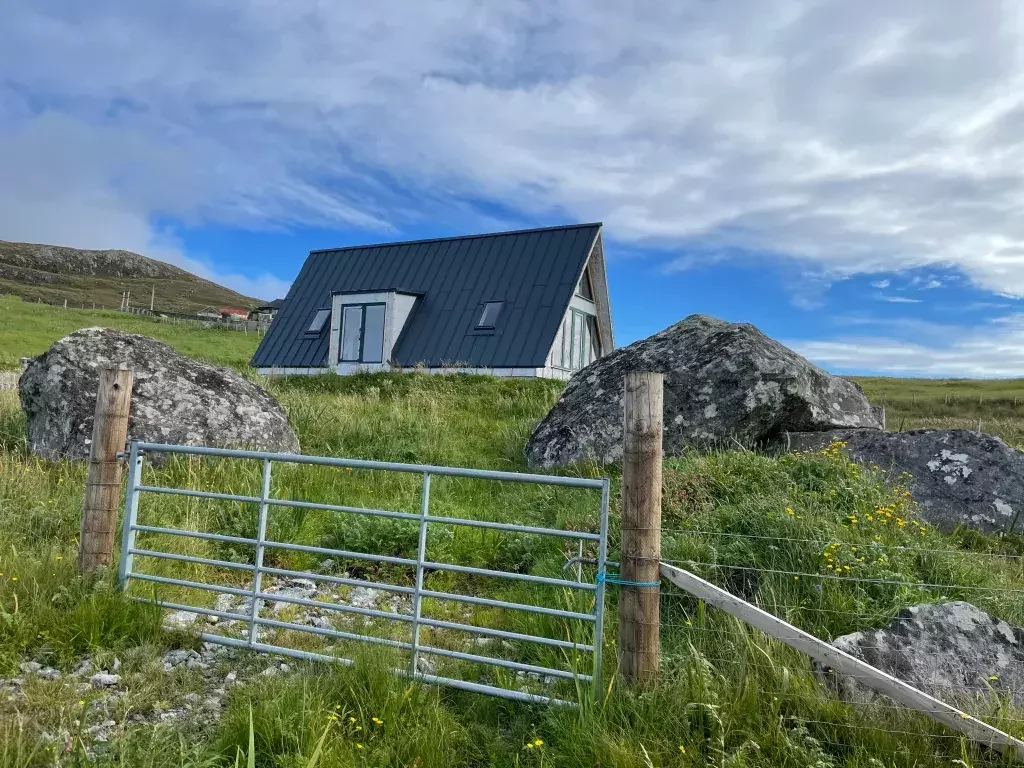 A modern A-frame house with a dark metal roof sits in a grassy, boulder-strewn field behind a metal gate under a bright, cloudy sky.
