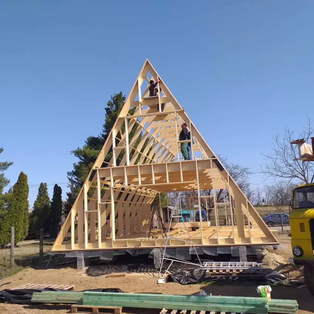 Two workers are pictured atop the wooden structural frame of an A-frame house under construction on a sunny, clear day.