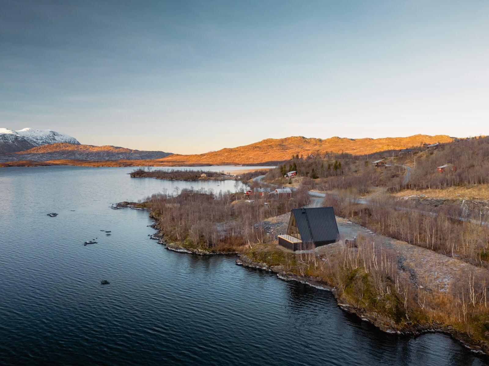 An expansive aerial view of a black A-frame cabin perched on a rocky shoreline, overlooking a vast lake and rolling golden hills under a clear twilight sky.