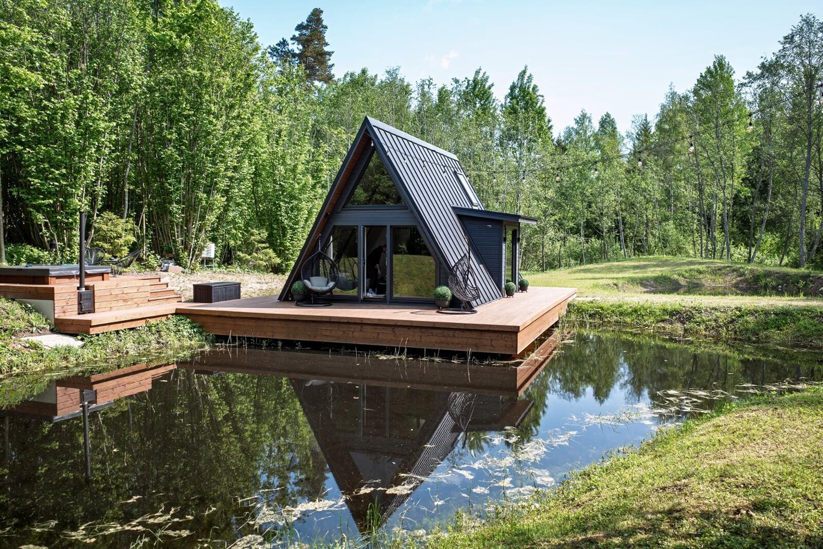 A modern black A-frame cabin sits on an expansive wooden deck that extends over a calm pond, surrounded by lush green trees and featuring a hot tub on a multi-level side platform.
