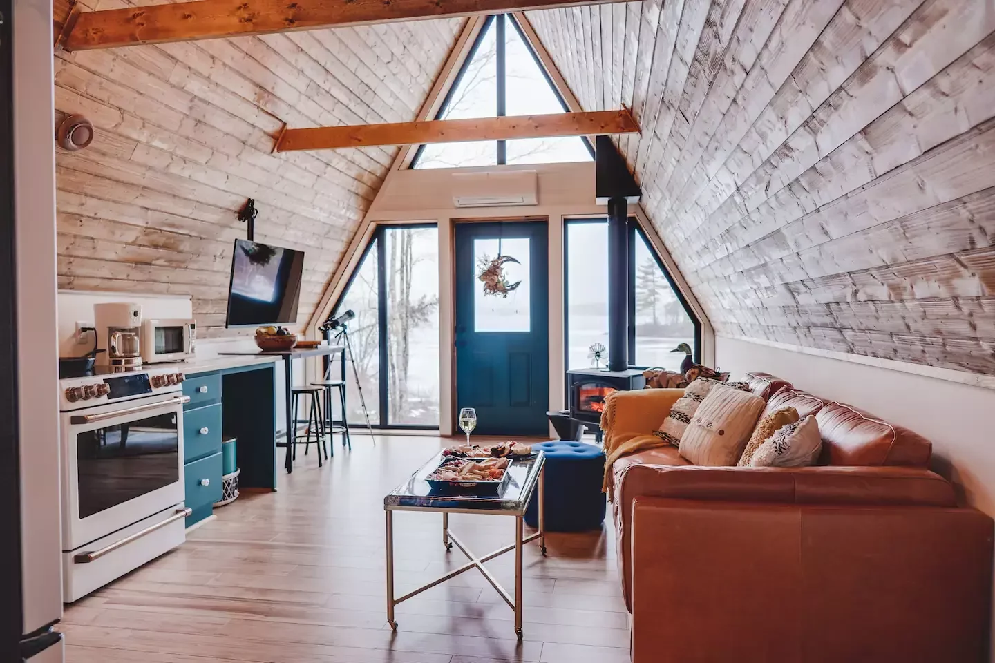 The interior of a cozy A-frame cabin featuring light wood-paneled walls, a tan leather sofa, and an open-concept kitchen leading toward a large window and dark blue front door.