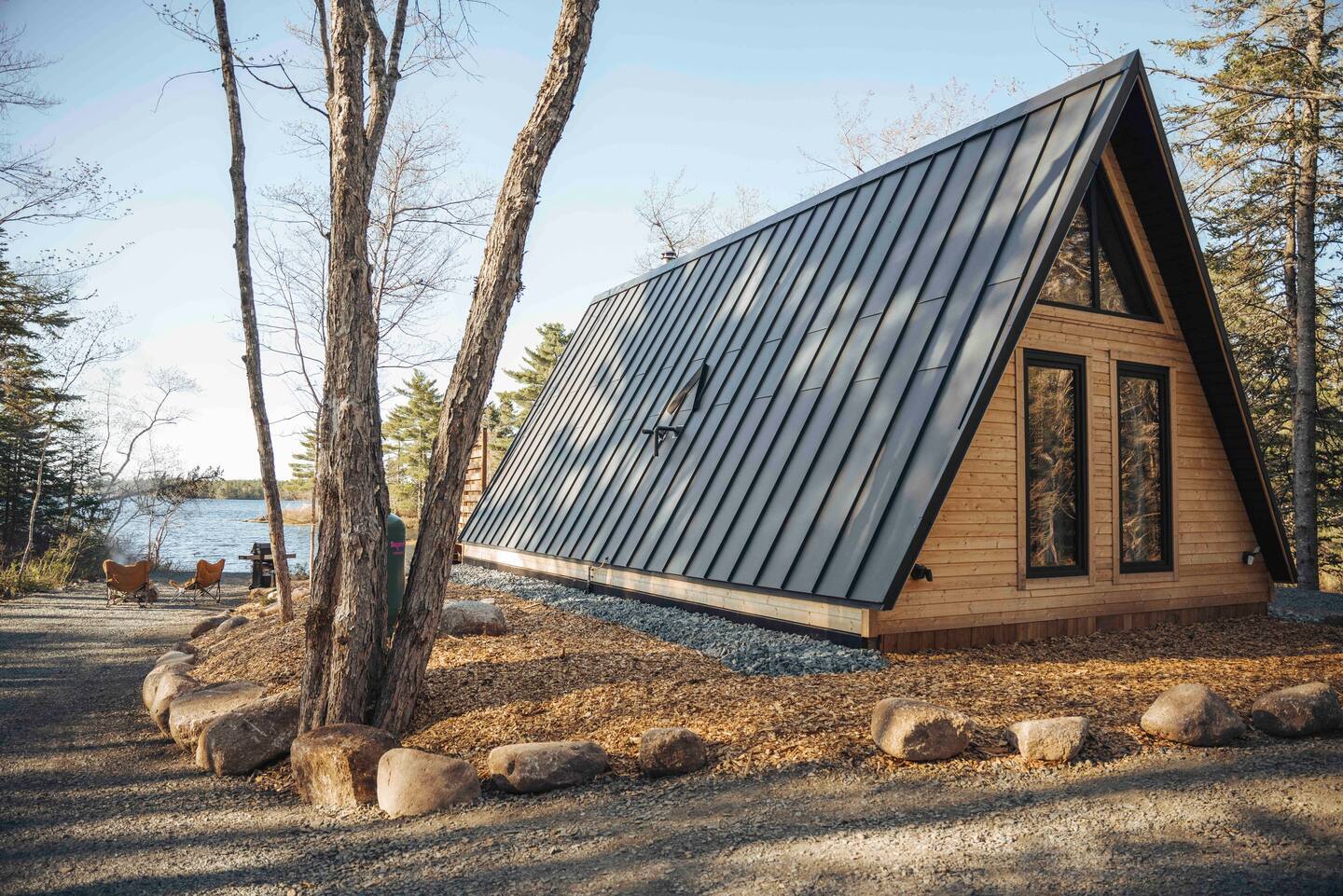 A side-angle view of a wooden A-frame cabin with a large black standing-seam metal roof, situated on a gravel lot near a calm lake.