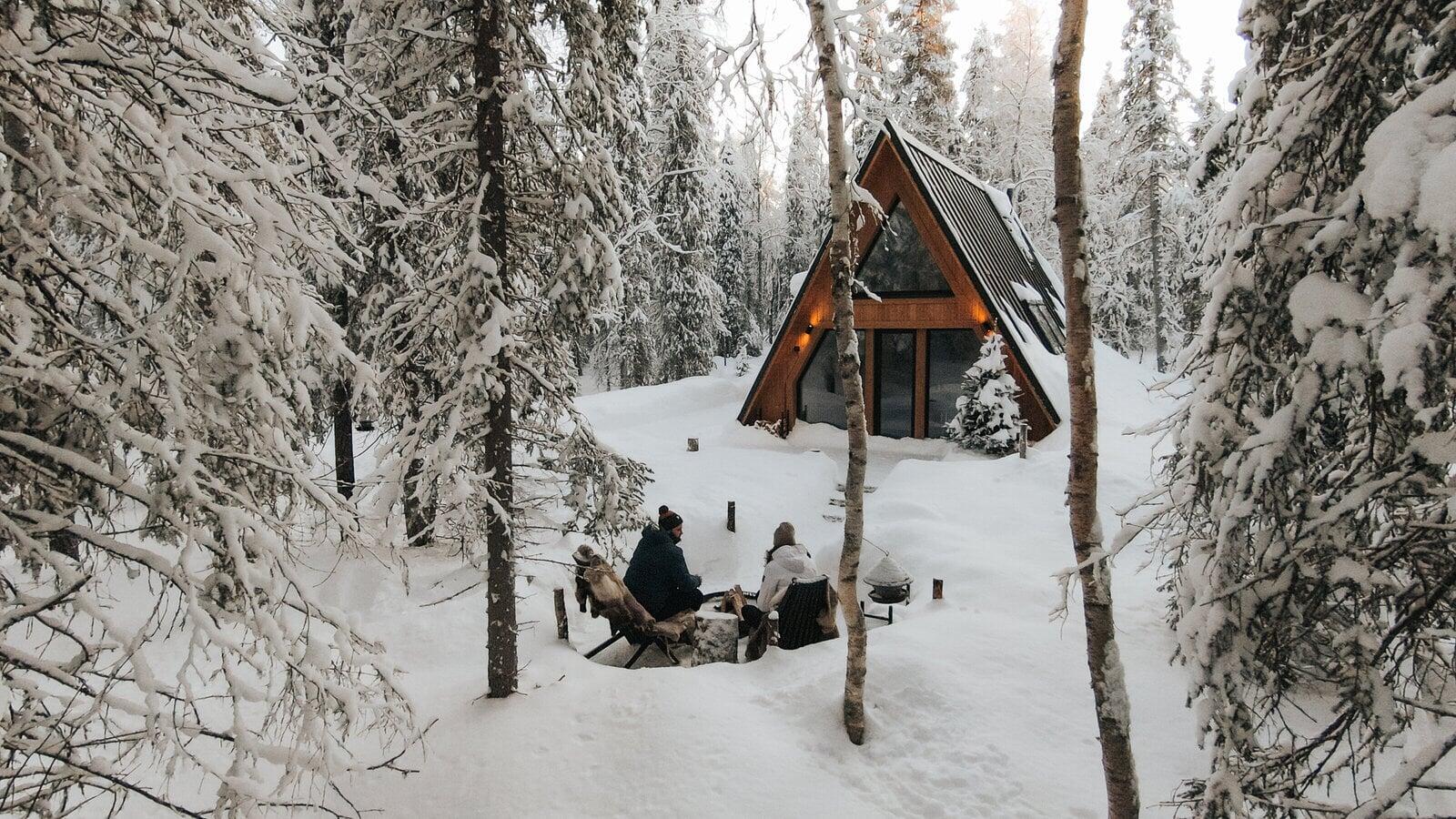 Two people sit by an outdoor fire in a heavy winter landscape, framed by snow-dusted trees with a warm-lit wooden A-frame cabin in the background.