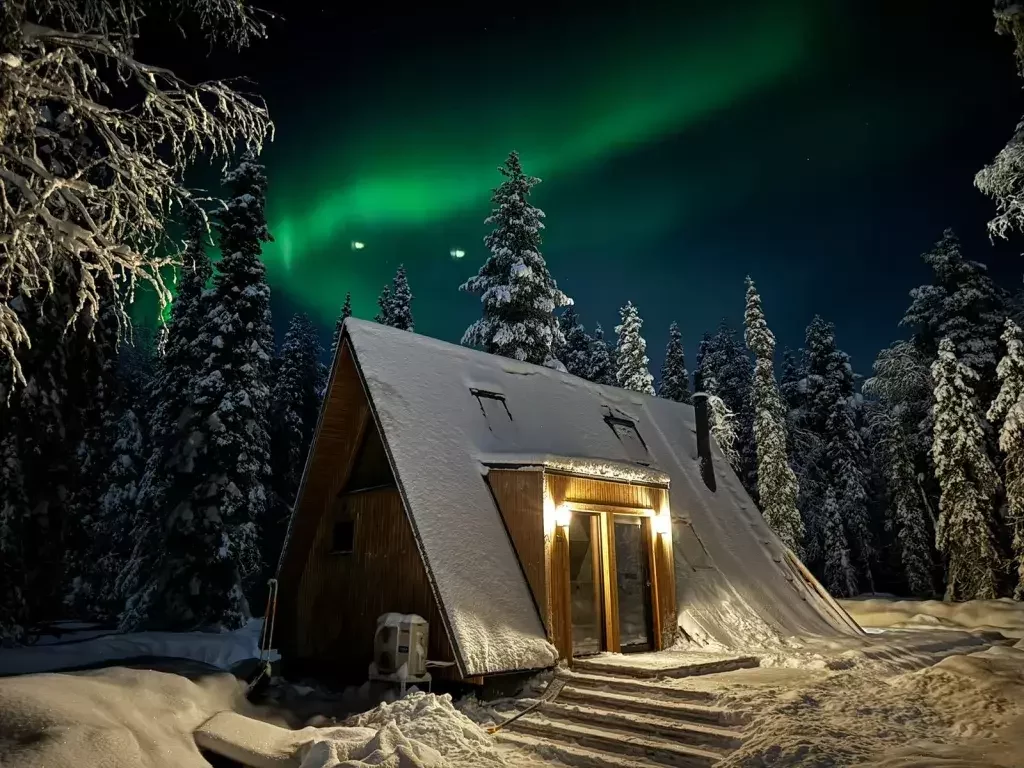 A glowing wooden A-frame house sits in a deep winter landscape under a vibrant green aurora borealis, surrounded by snow-covered evergreen trees.